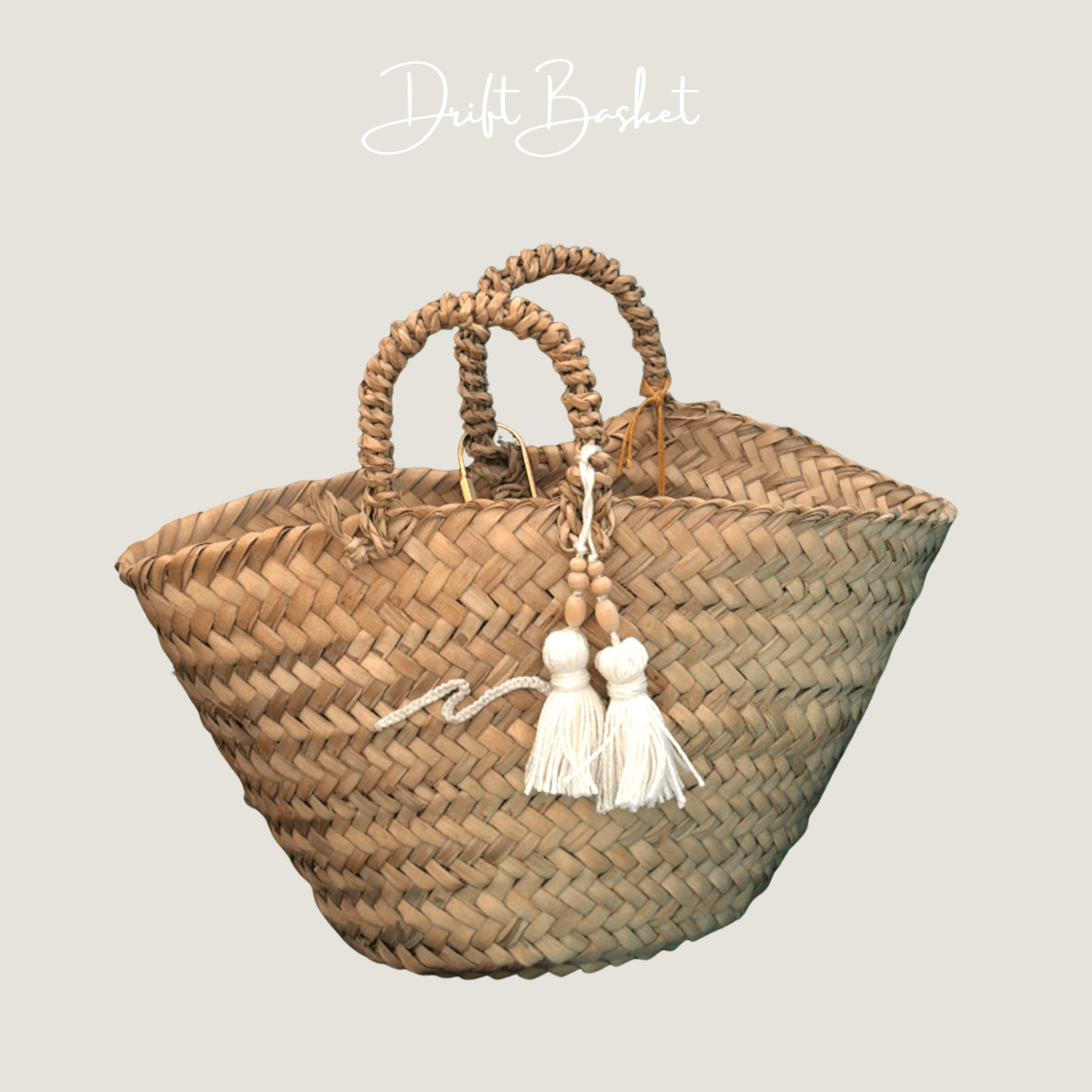 A natural woven straw tote bag with two rolled top handles and two white tassels with wooden beads hanging from the front, shown on a plain light gray background with the text 'Drift Baslet' at the top.