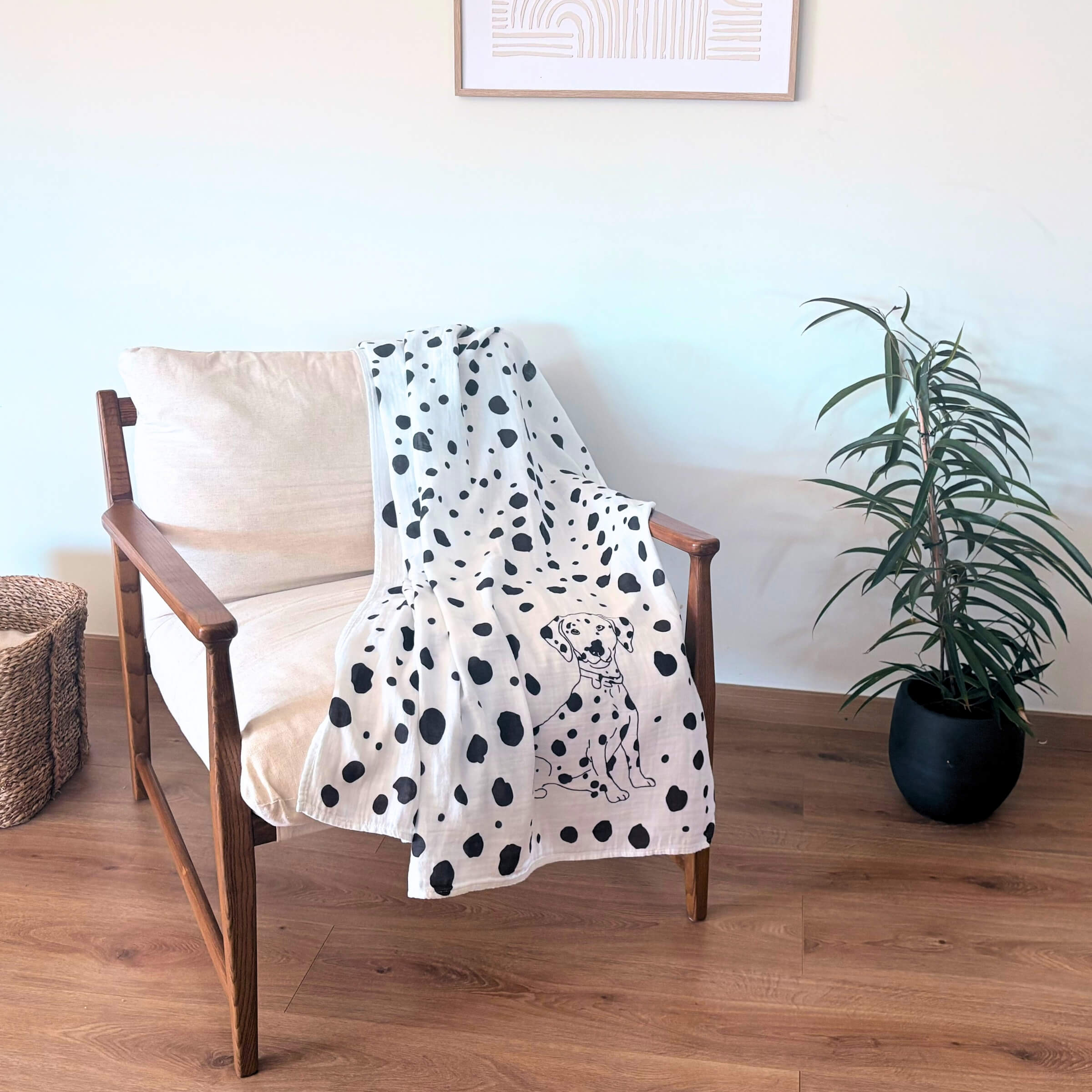 Modern chair with a Allure en Noir Dalmatian patterned Large Square blanket draped over it, next to a plant on a wooden floor.