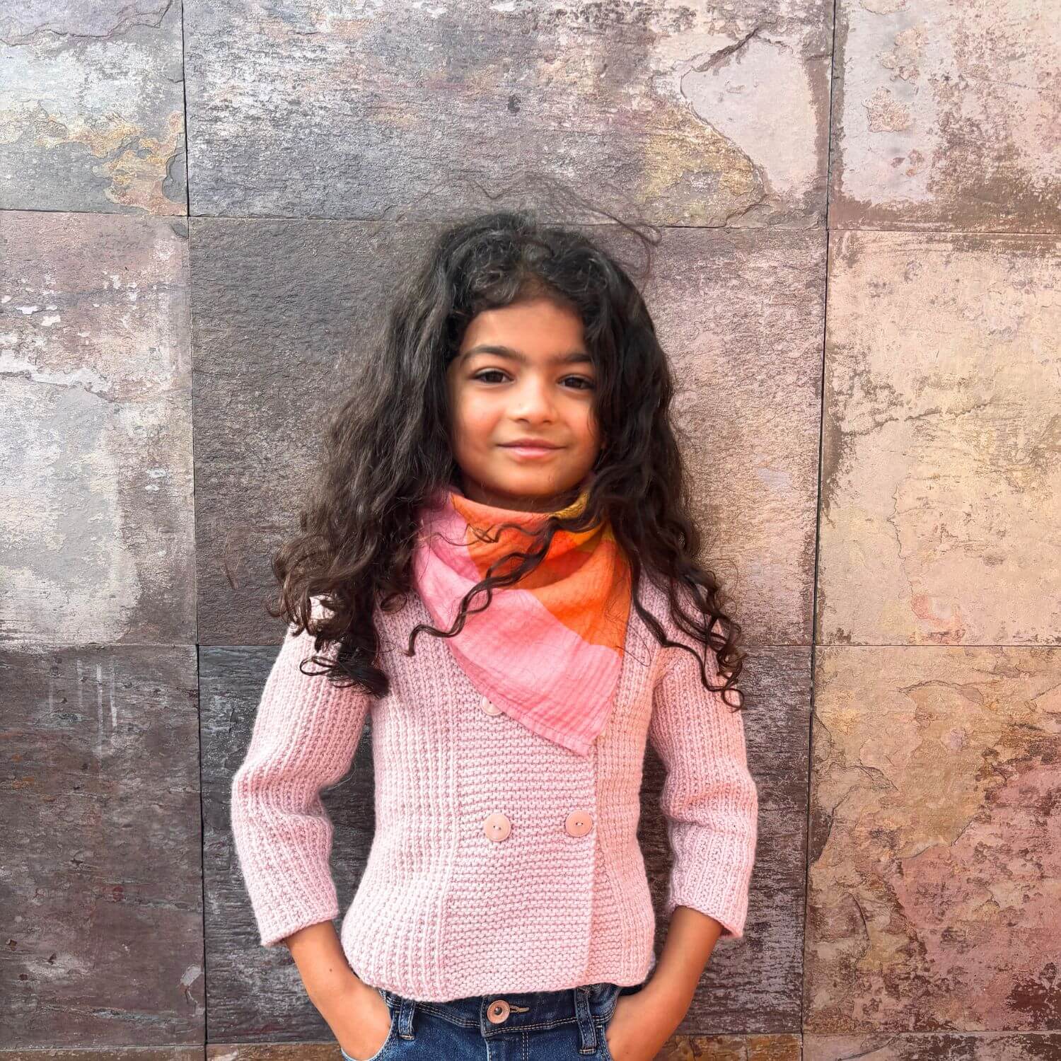 Young girl wearing a pink sweater and colorful scarf against a textured wall.