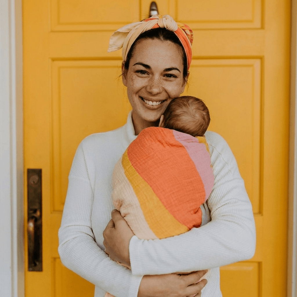 Woman holding a baby wrapped in a colorful blanket in front of a yellow door.