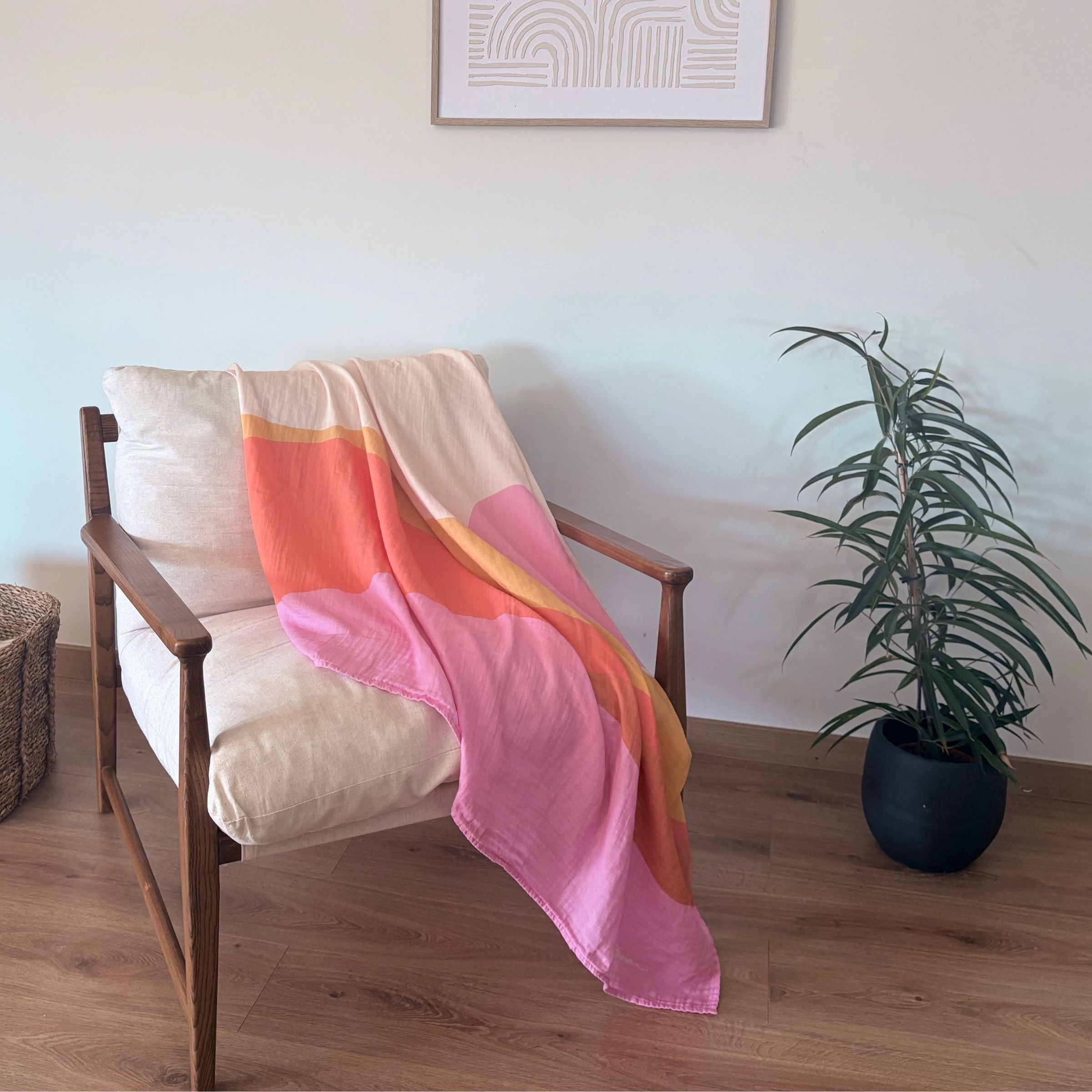 The Large Square Indra as Sofa throw towel cotton blanket, draped over a modern wooden chair in a living room with a palm tree plant and framed picture on the wall.