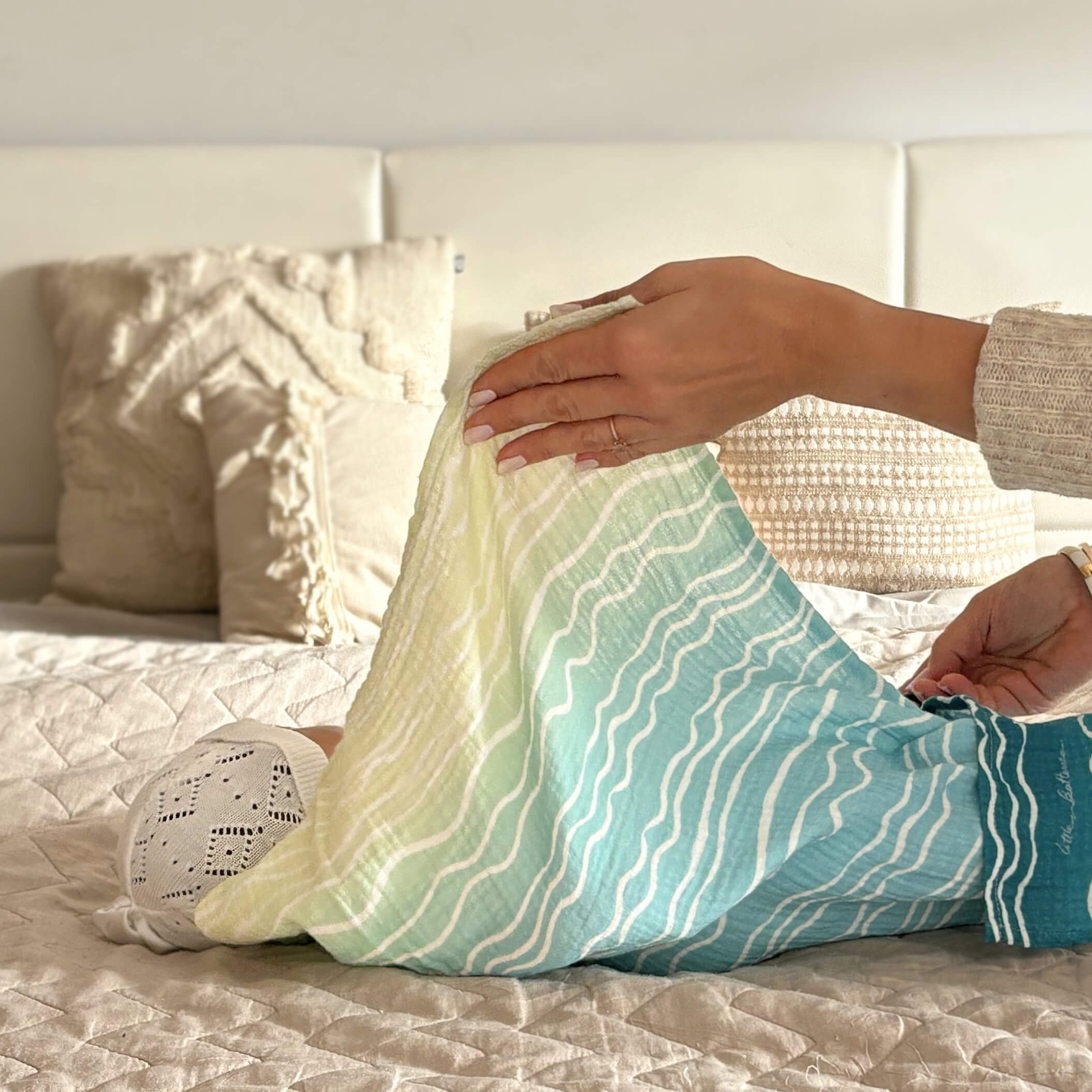 Hands gently arranging the Brisa do Mar Small Square muslin on a bed, showing softness and ease during everyday use.