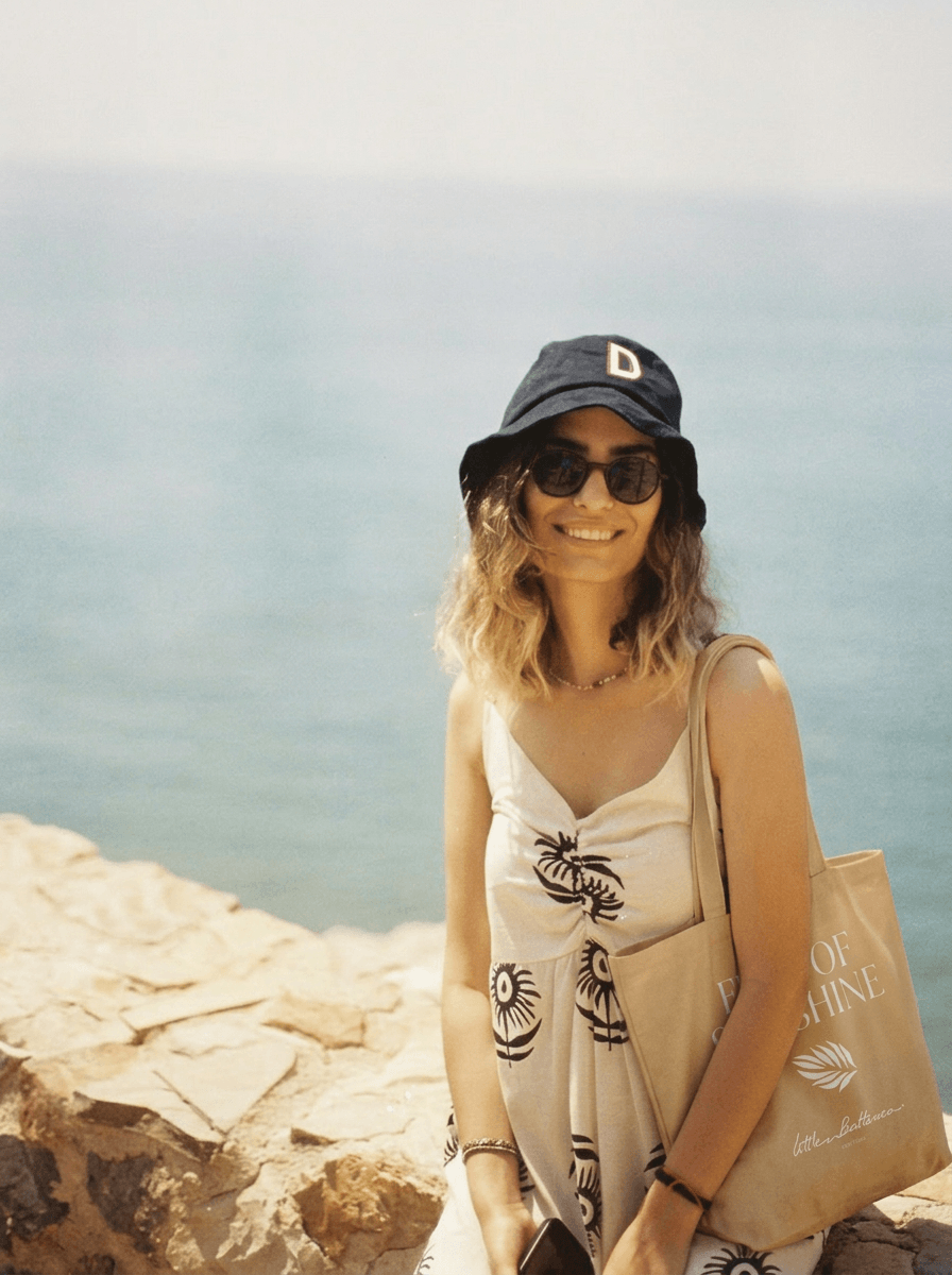 Woman standing on a rocky beach holding a beige bag with text, wearing a black cap and sunglasses.