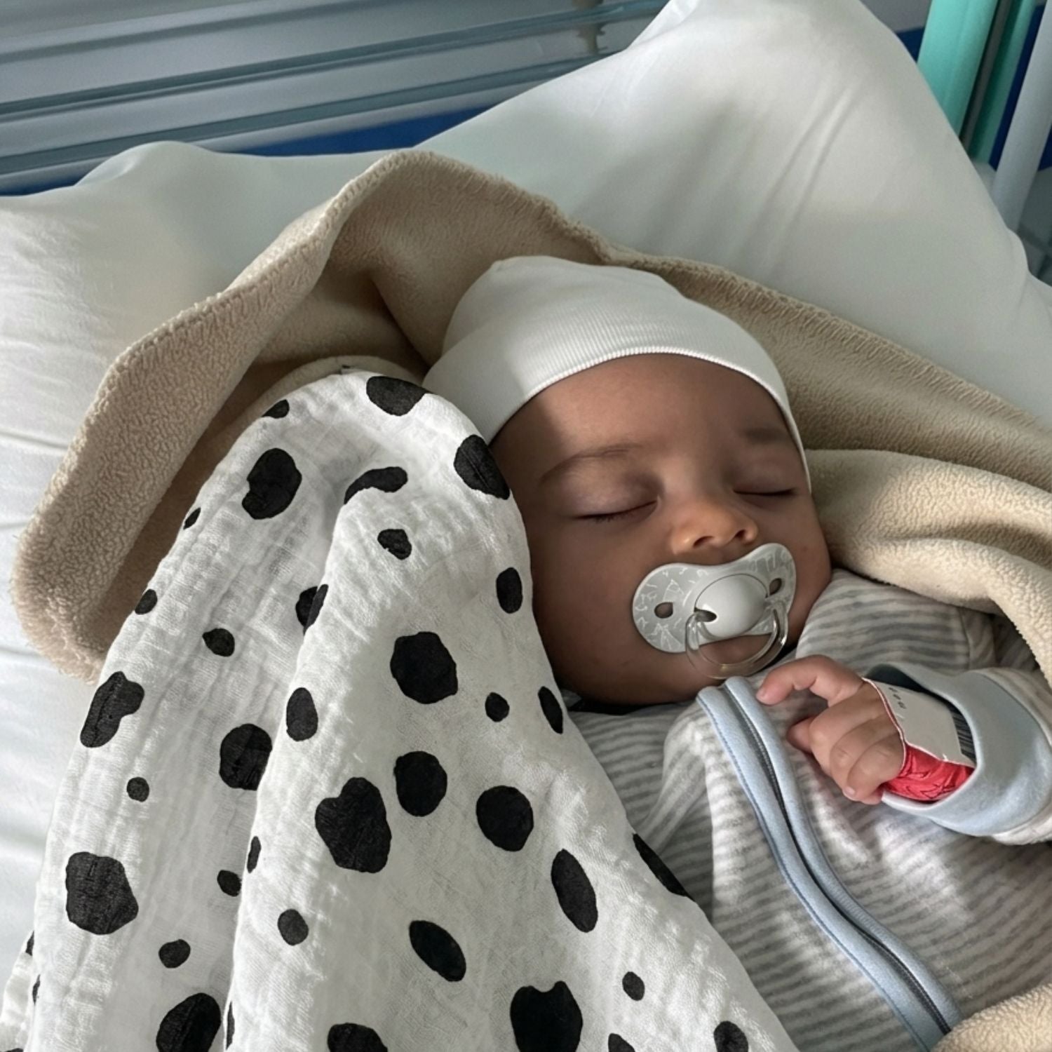 Baby sleeping with a pacifier, wrapped in Allure en noir Large Square muslin blanket and wearing a white hat.